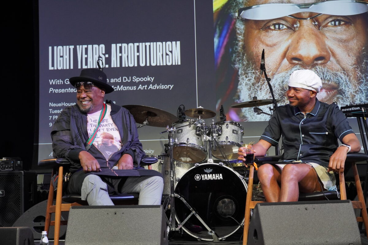 George Clinton and DJ Spooky watch the audience smiling while seated on a stage in front of drums and a screen reading in the background 