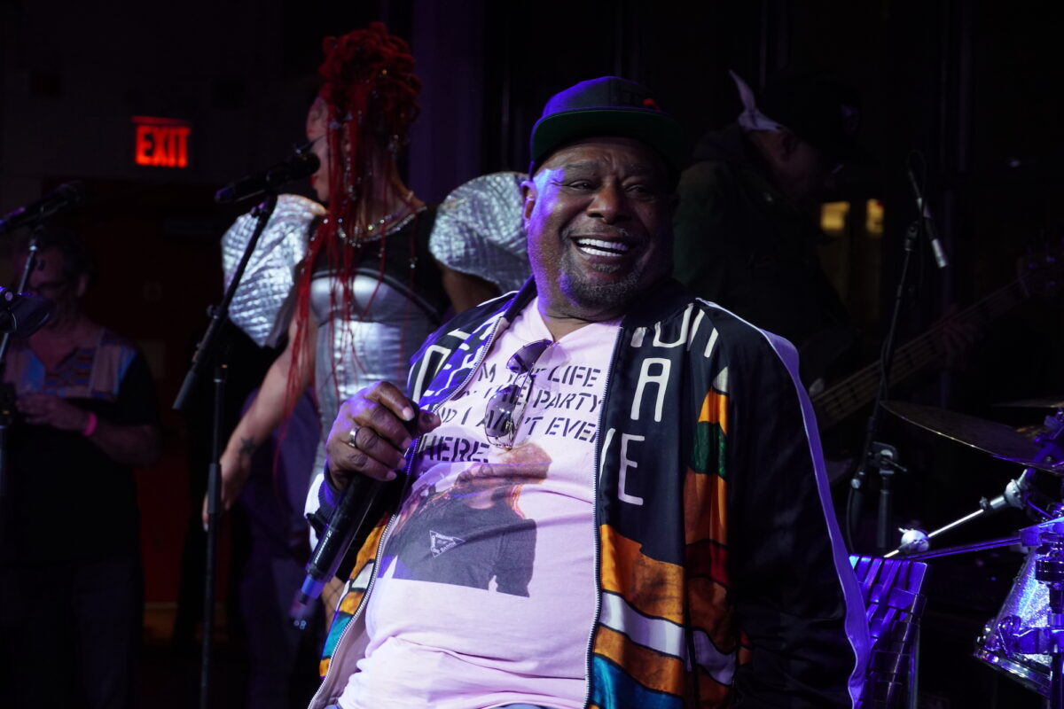 George Clinton holds a microphone and smiles as he stands on a stage