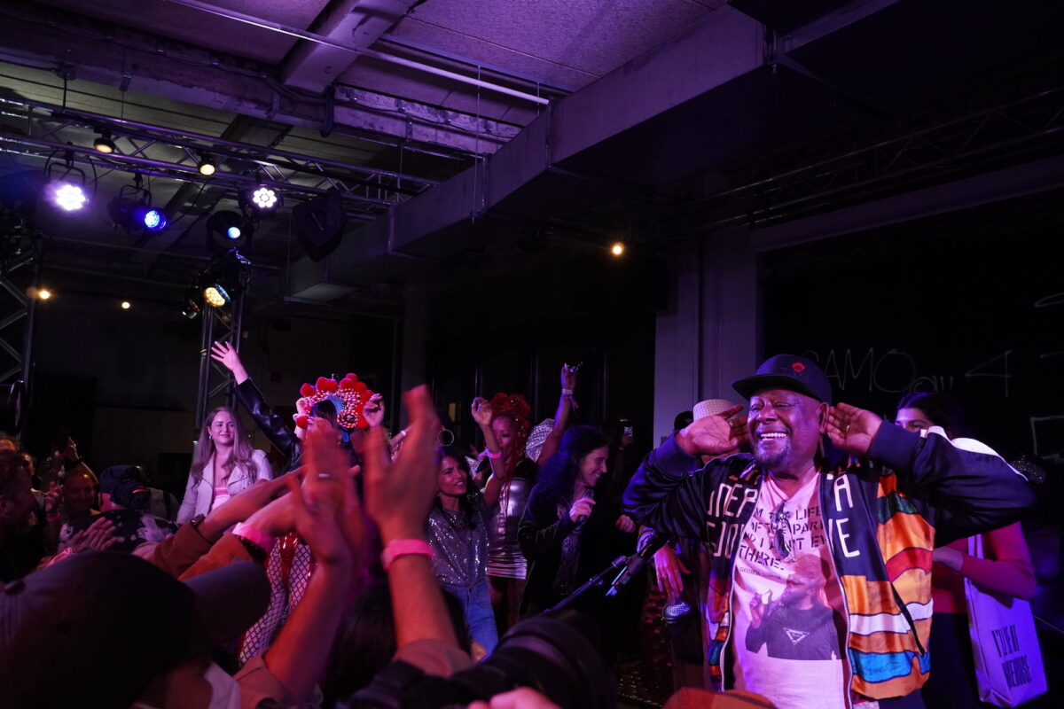 George Clinton joyfully pushes his ears forward so as to hear the audience, as fans dance and clap along on and off stage