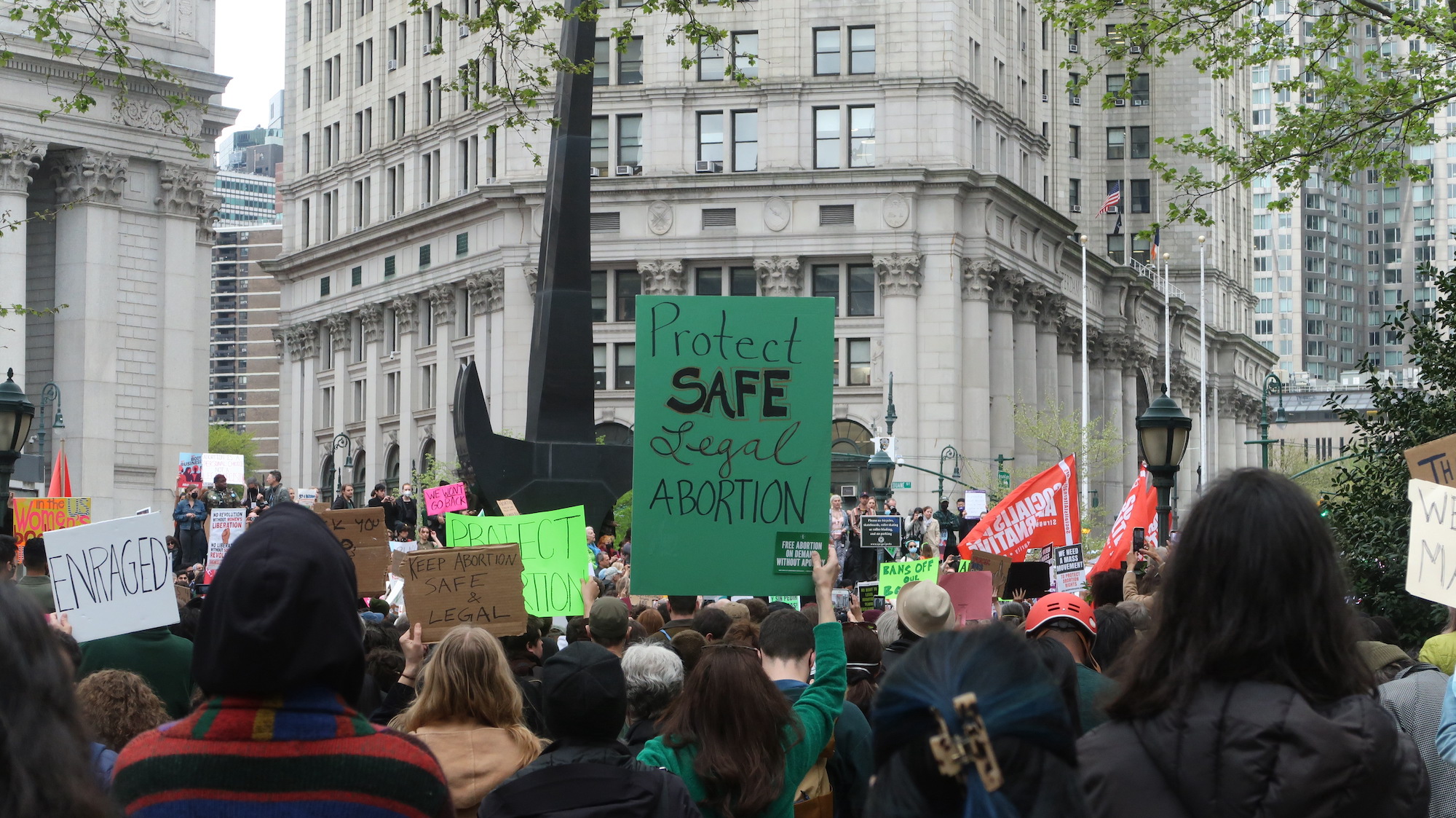 A crowd of protesters faces an elevated group of speakers. A green sign in the center reads “Protect safe legal abortion”