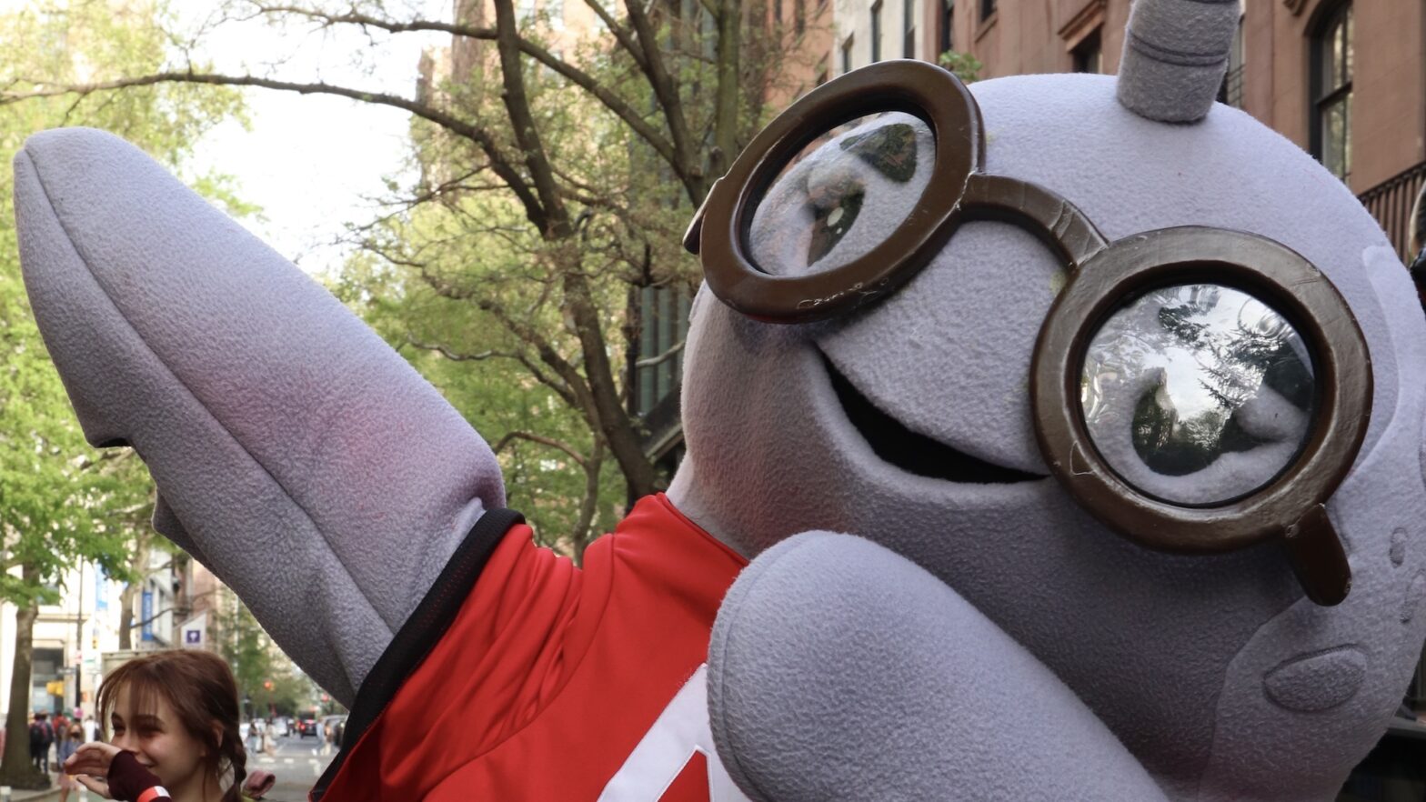 A person dressed in a New School mascot narwhal costume wears a New School shirt and dabs on the street.
