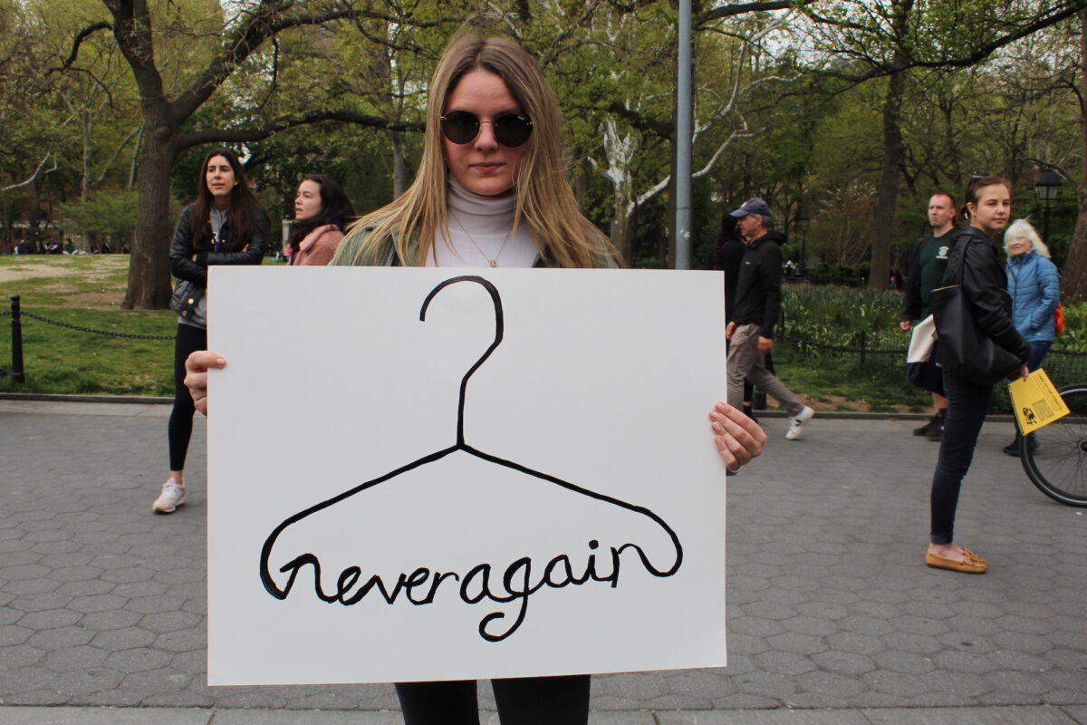 A woman  holds up a poster with a drawing of a coat hanger, which has been twisted to read “never again”