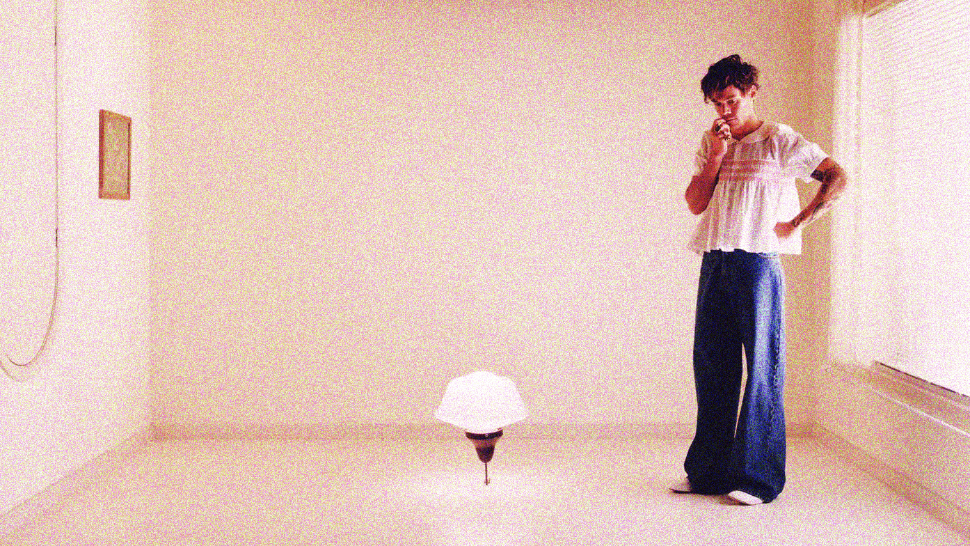 Harry styles stands in an upside down white room with a pendant light hanging from the center. He wears flare jeans and a delicate white blouse, and stands with one hand on his hip and one up to his mouth. He looks downwards in a melancholy way, as though thinking