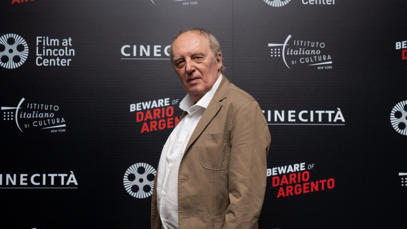 Dario Argento stands in front of a poster board with the logos of Italian film organizations.
