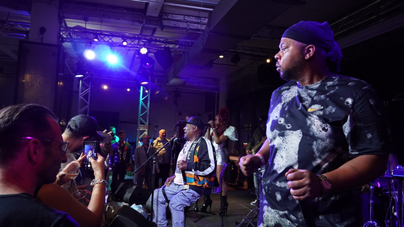 George Clinton, sitting on stage at a performance in NYC with two other vocalists and an enthusiastic crowd.