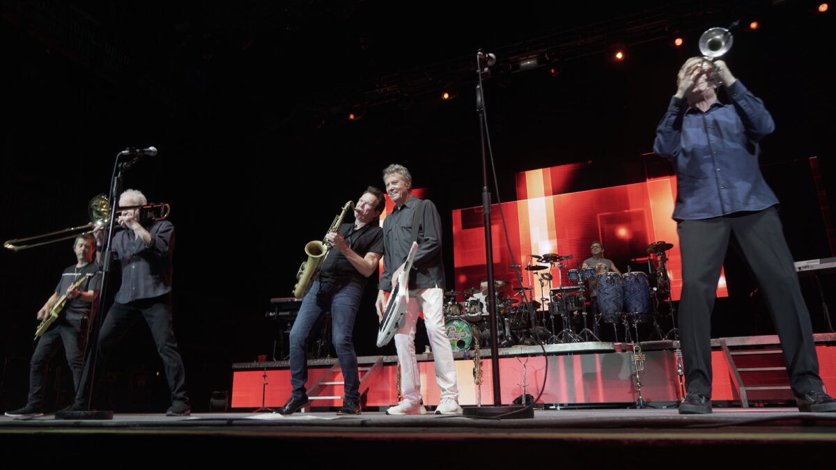 Robert Lamm plays the keytar alongside other band members playing the guitar, tuba, trumpet, guitar and drums, backdropped on stage by an abstract red screen of color.