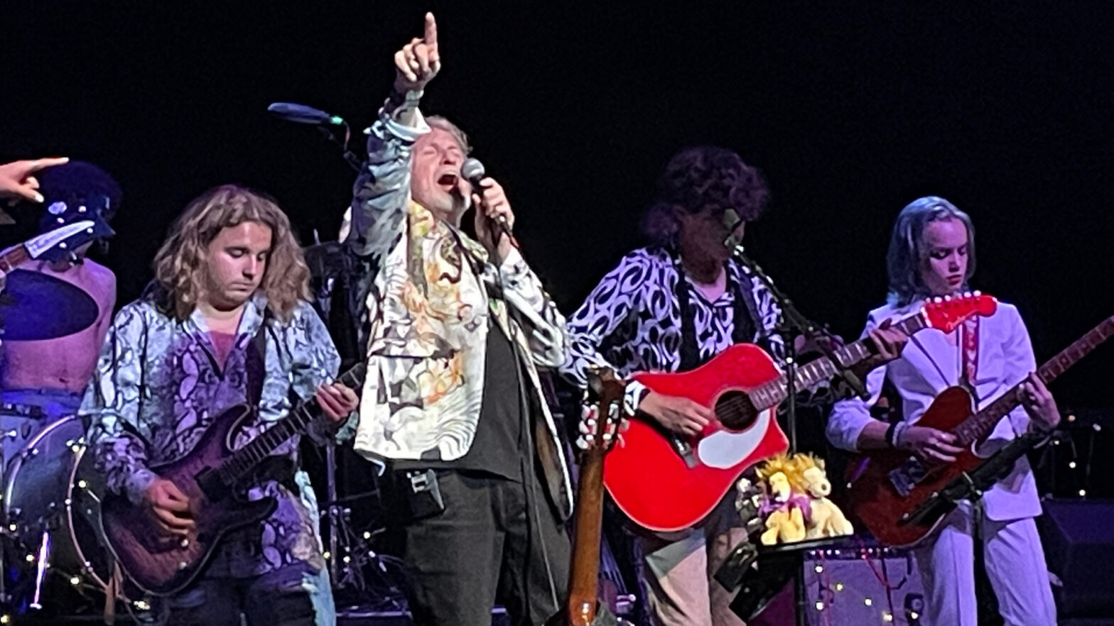 Jon Anderson singing onstage with teenage students on guitars, backdropped by a dark stage.