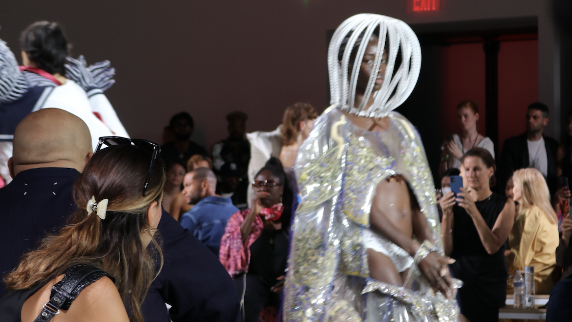 A crowd watches a model walking in a white headpiece and gold vinyl dress.