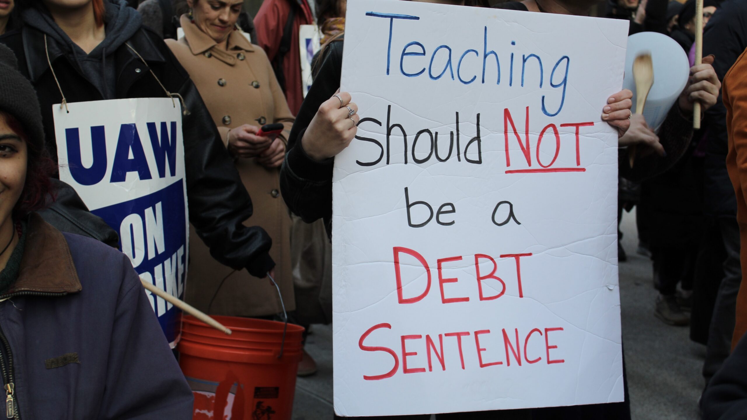 A sign held up by a protestor in front of The New School’s university center saying “Teaching should not be a debt sentence”.