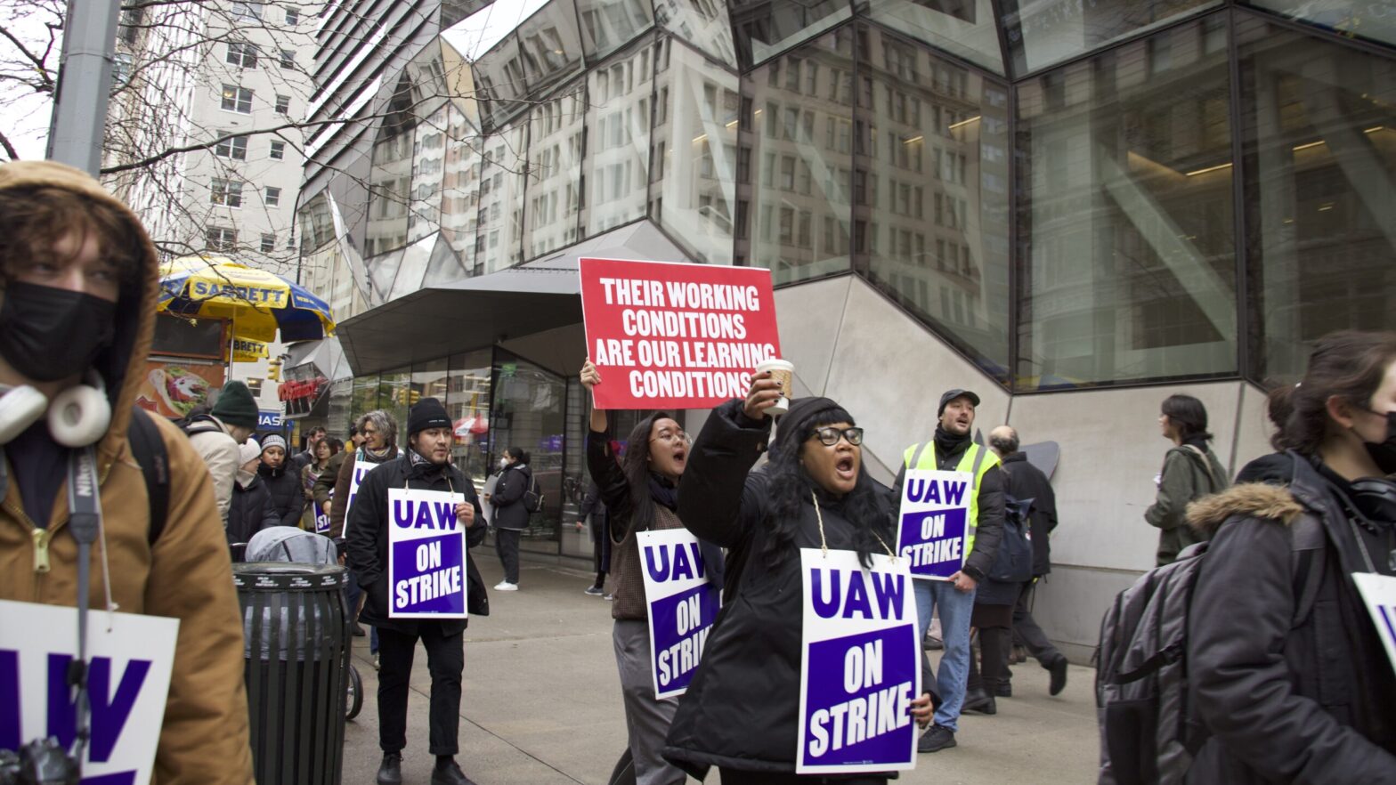 Protesters gather outside of The New School with signs there is one bright red sign that reads ‘Their working conditions are our learning conditions’ and many people with blue ‘UAW ON STRIKE’ signs.