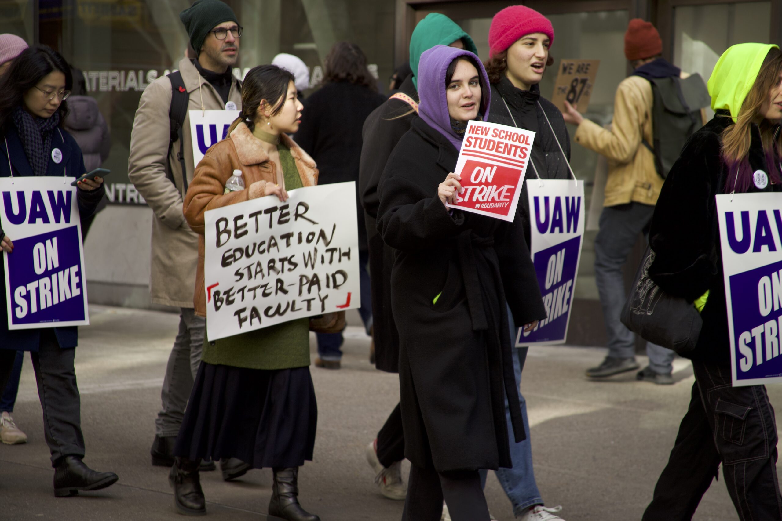 Students outside of The New School hold signs. One is white with black handwritten text that reads 'Better education starts with better-paid faculty" and other white sign with red text reads 'New School students on strike.' They are surrounded by people with blue signs that read UAW On Strike.