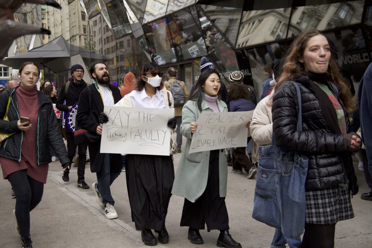 Two students with white signs with handwritten text that say ‘Pay the fucking faculty’ and “Teachers deserve a livable wage. They are in line picketing with other people marching and yelling. 