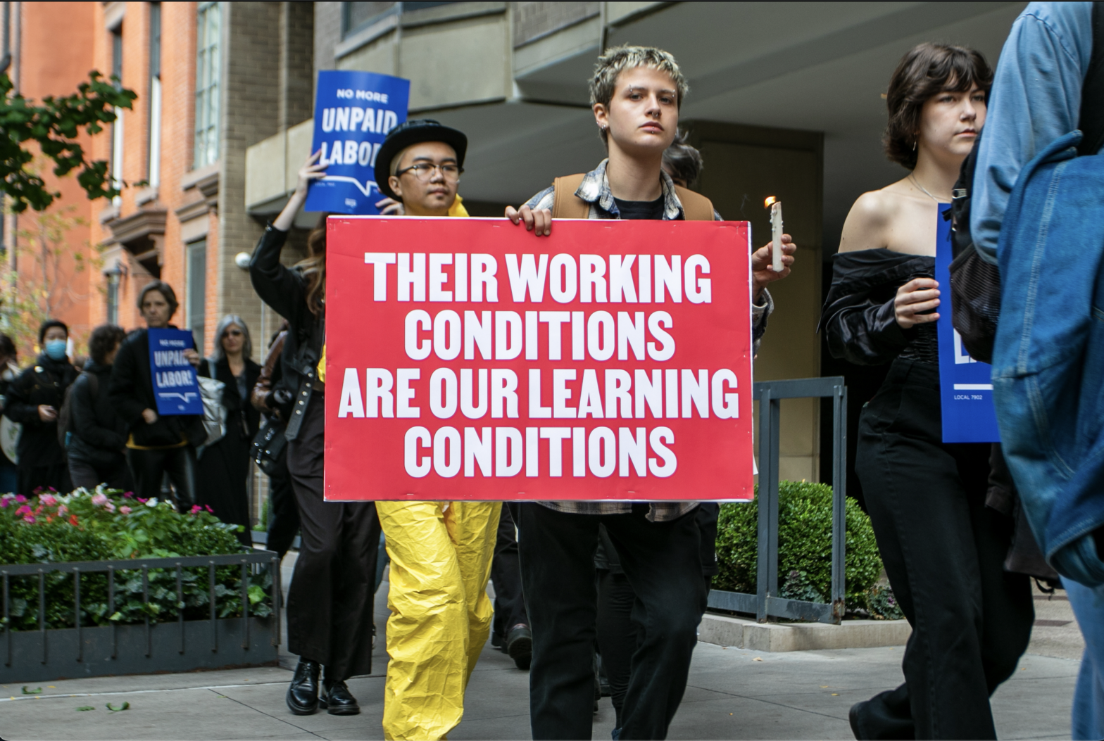 Students march in solidarity with The New School’s part-time faculty union. One student holds a sign in one hand that reads, “THEIR WORKING CONDITIONS ARE OUR LEARNING CONDITIONS,” and in the other holds a melting candle.