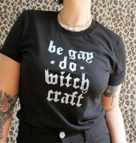 A person wearing a t-shirt created by Midge that says “be gay do witch craft”. 