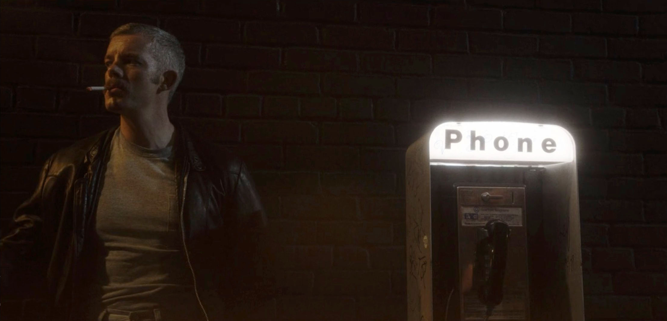 A man in a leather jacket smoking a cigarette next to a pay phone.