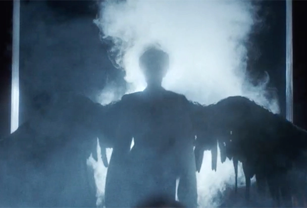 A shadowy human figure with large  angel wings and smoke behind it. 