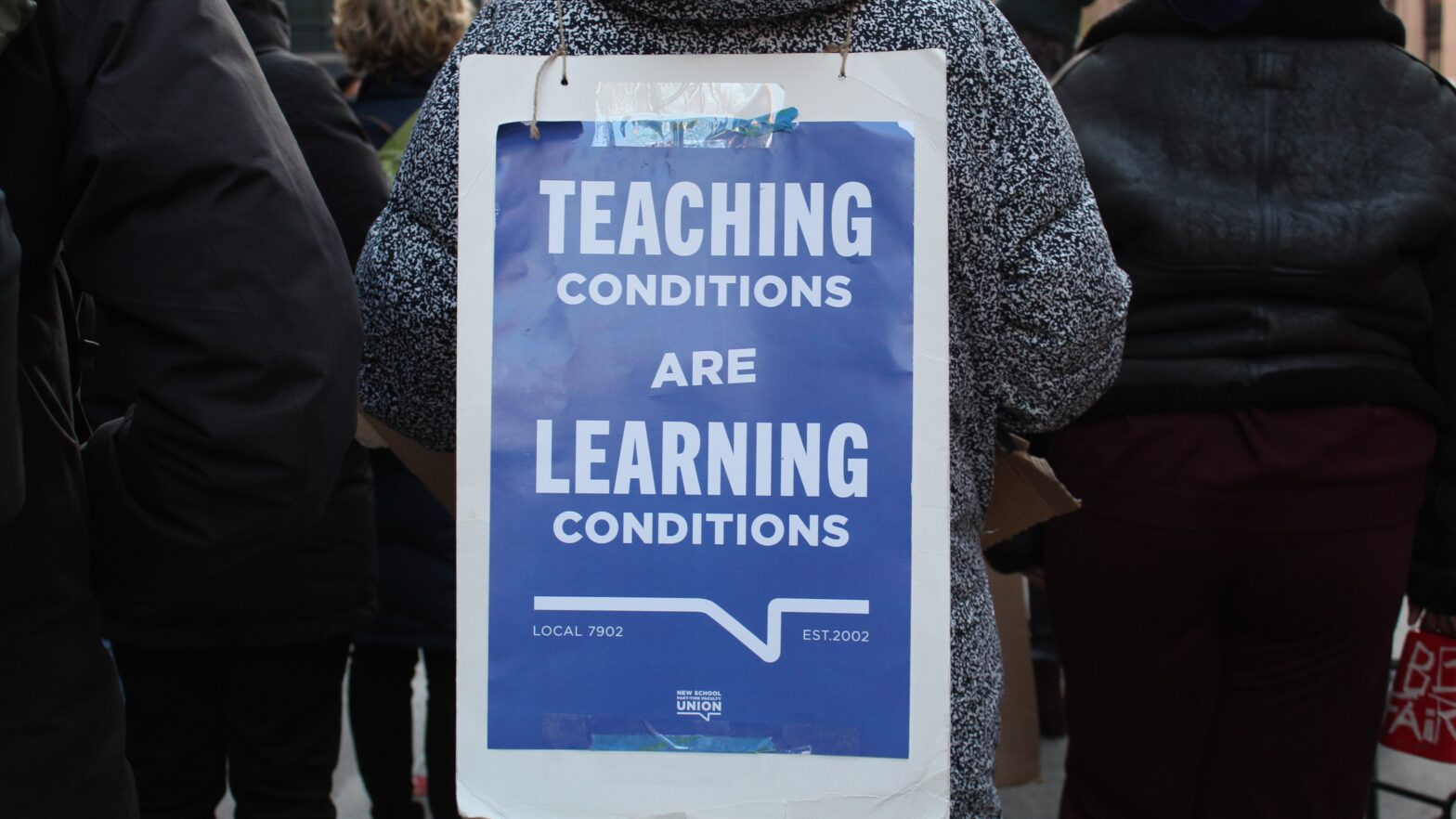 A blue and white sign reading “Teaching Conditions Are Learning Conditions” placed on the back of a protester.