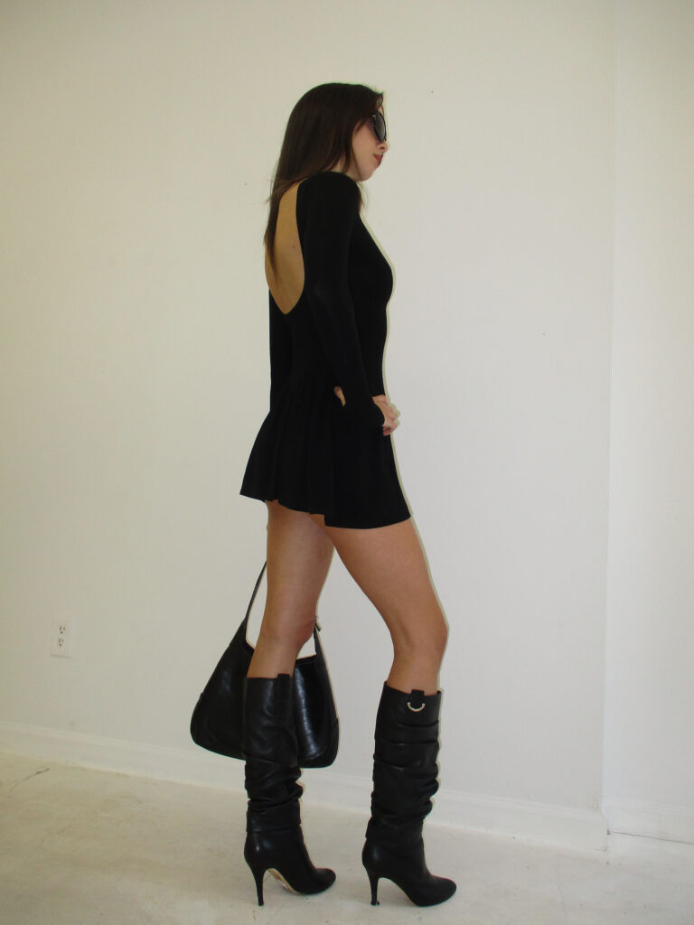 Model stands against wall, with shoulder turned to the camera. She wears black knee-high boots, a black mini skirt, and a long sleeved black shirt. She carries a black bag in her right hand, and wears big black sunglasses. 