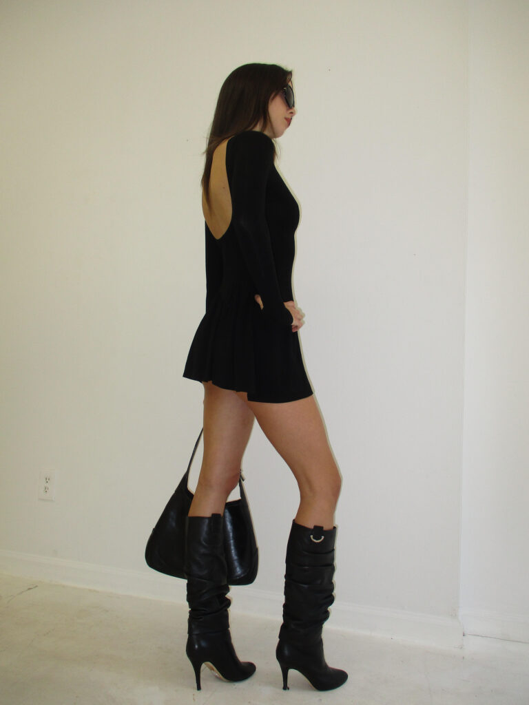 Model stands against wall, with shoulder turned to the camera. She wears black knee-high boots, a black mini skirt, and a long sleeved black shirt. She carries a black bag in her right hand, and wears big black sunglasses.