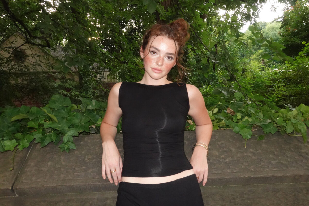 Red headed model leans against a ledge, in front of foliage. She wears a black sleeveless boat neck tank top, and a black mini skirt.
