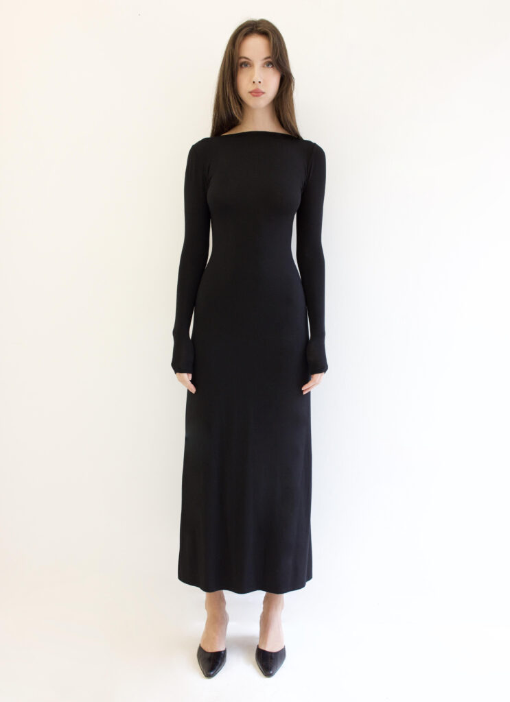 Model stands in front of a white backdrop, wearing a long-sleeved, floor length black dress, and black pointed kitten heels.