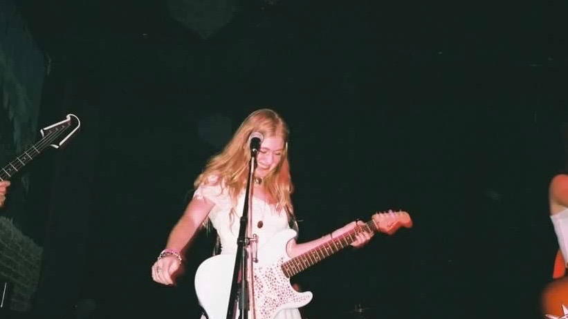 True Jackson stand on stage at a performance, wearing a white dress and playing a white guitar. 