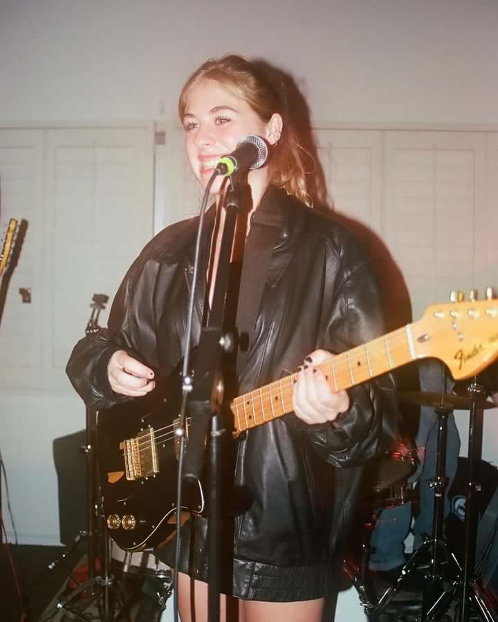 True Jackson stands in front of a microphone, performing at a concert. She wears a skit and black leather jacket, playing a black guitar.
