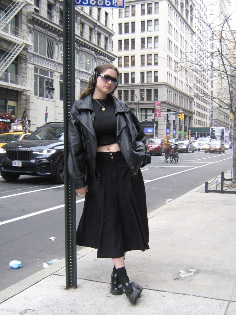 fashion student wearing all black with headphones and sunglasses standing under fifth avenue sign.