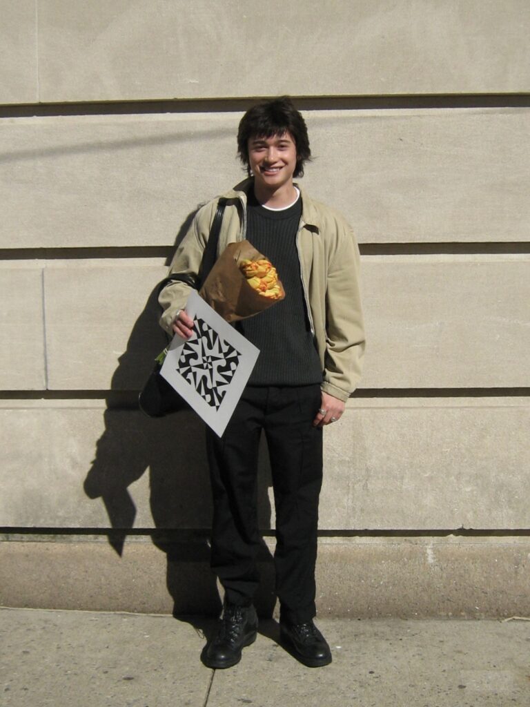 student holding tulips and art project wearing a black outfit with a beige jacket standing against beige wall.