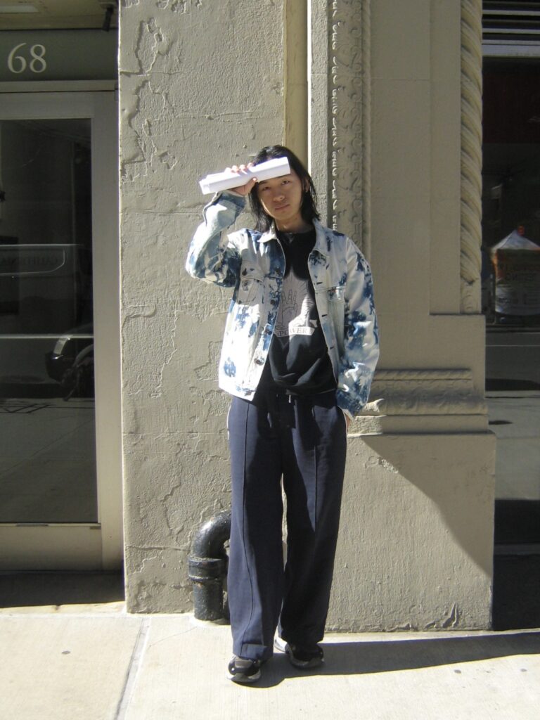 student in sneakers, slacks, a crewneck sweatshirt, and denim jacket standing against building on 5th avenue