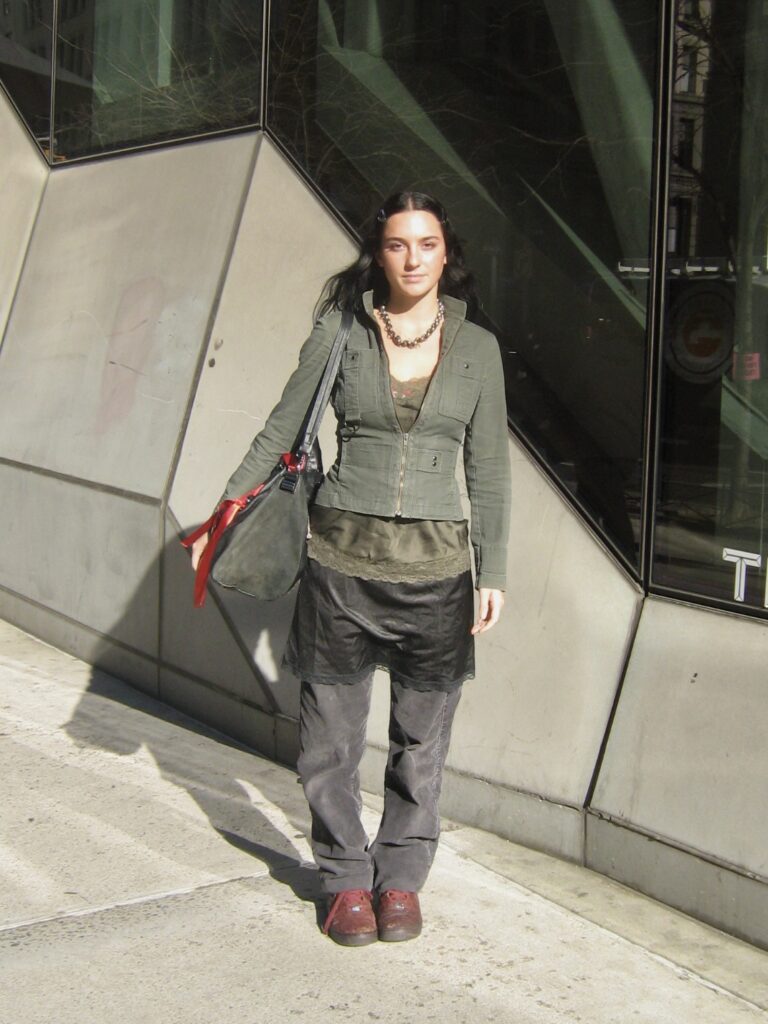 Fashion student in a green jacket, and a black skirt layered over pants standing on 5th avenue.