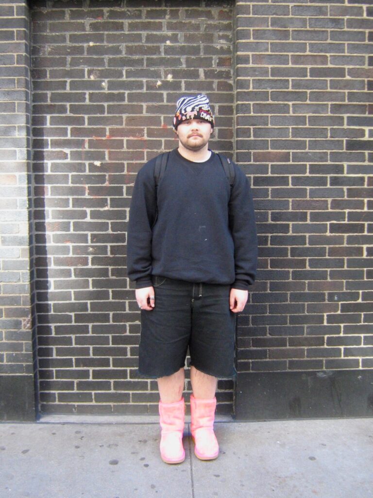Student wearing black outfit with a beanie and pink uggs standing against brick wall.

