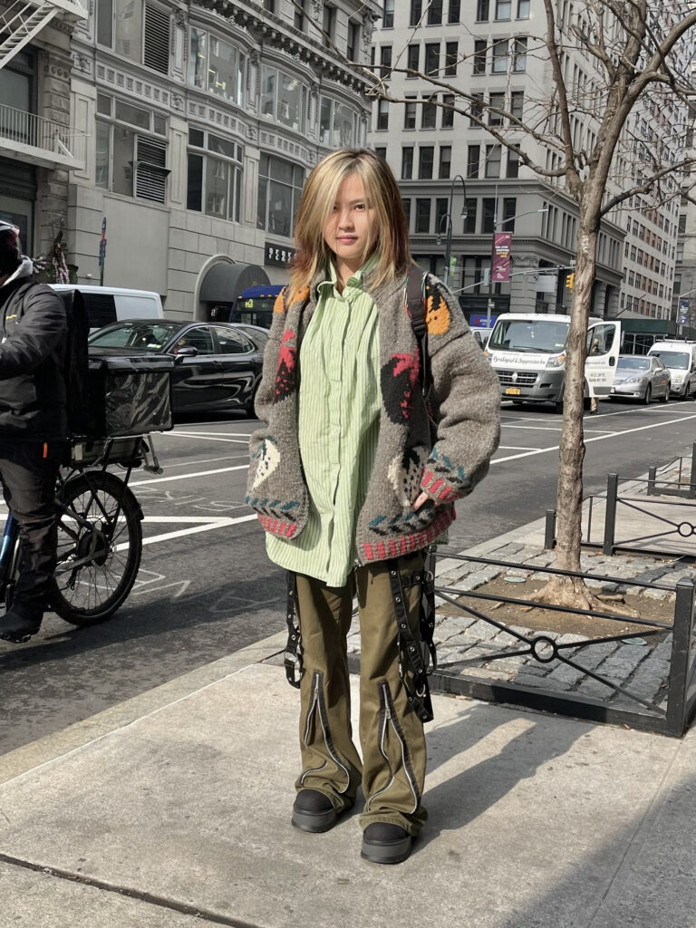 Parsons student wears a knit strawberry cardigan, a green button up shirt, and black and green cargo pants standing on fifth avenue after class.
