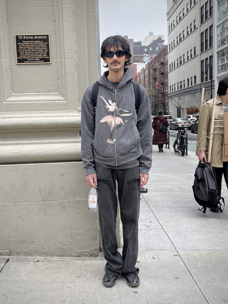 Parsons student is comfortable and incognito in black jeans, a gray graphic sweater, and black sunglasses while standing on the corner of fifth avenue.