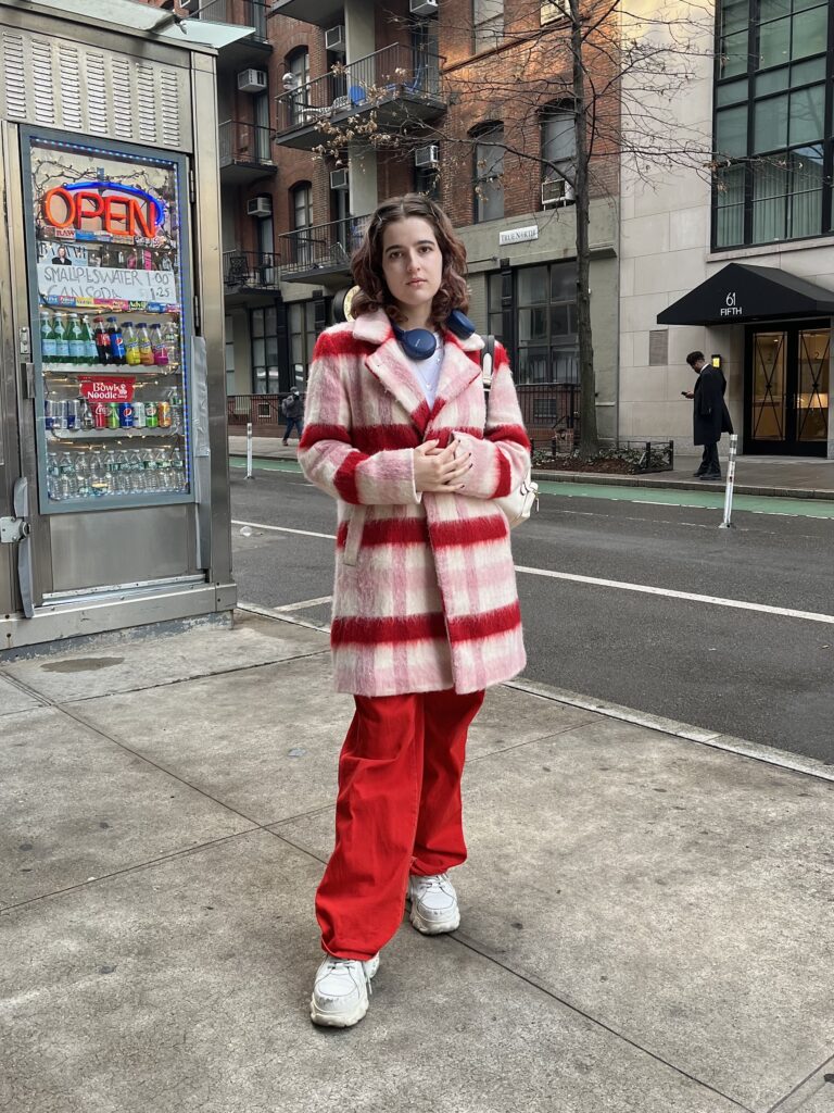 Drama student wears a plaid pink, red, and white peacoat with red pants and white sneakers standing on fifth avenue.