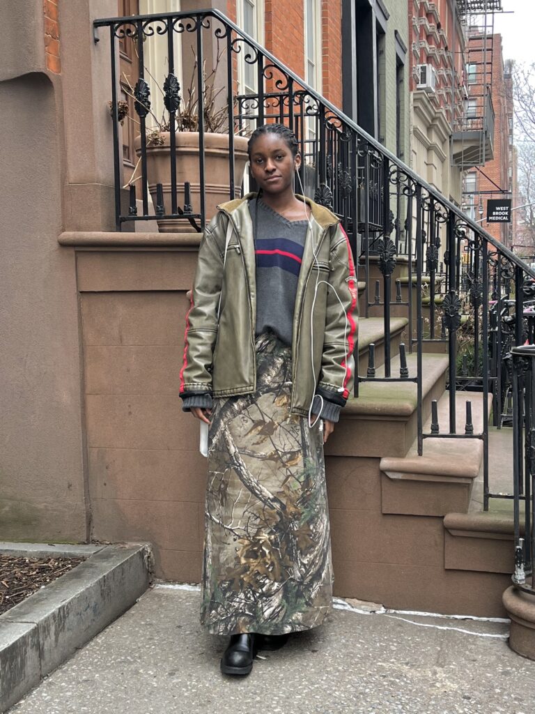 Art school student wears a camouflage maxi skirt, striped sweater, and green leather jacket while listening to music before classes.