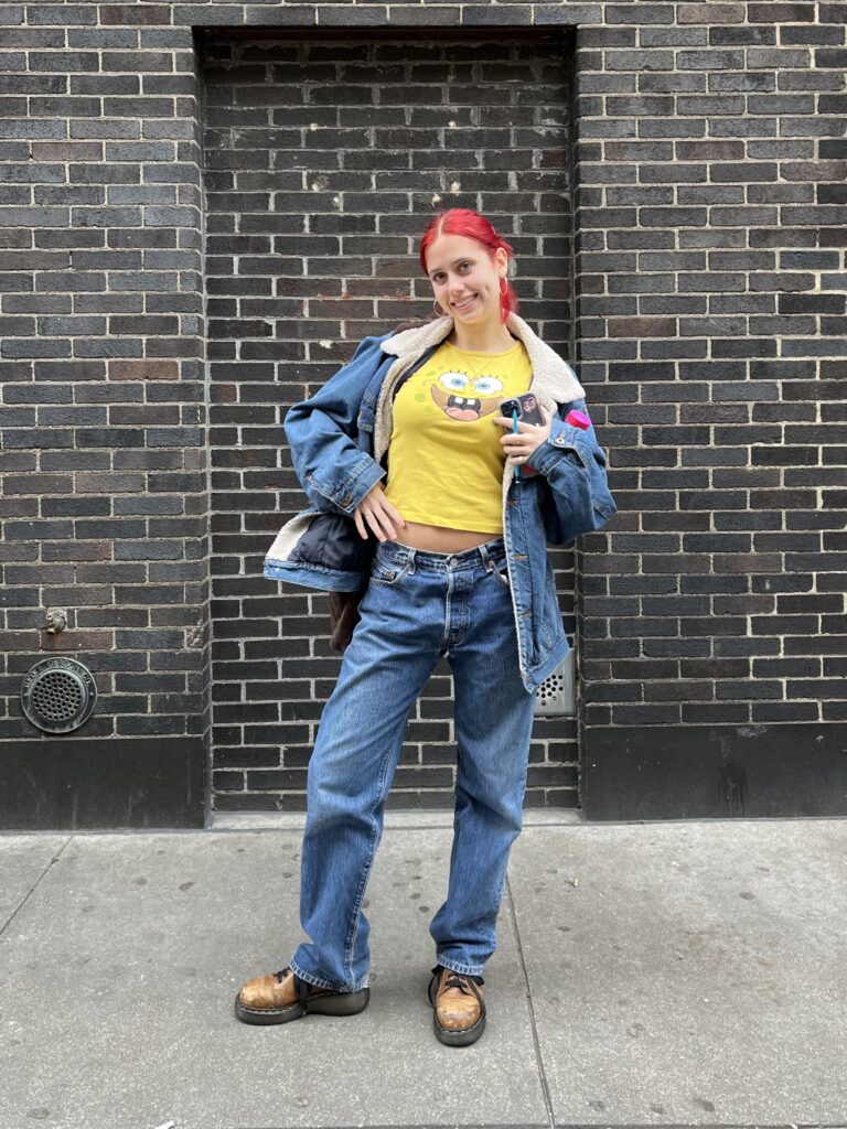 Art school student wears a matching denim jacket with baggy jeans and a yellow spongebob t-shirt with her bright red hair standing with a smile in front of a brick wall.