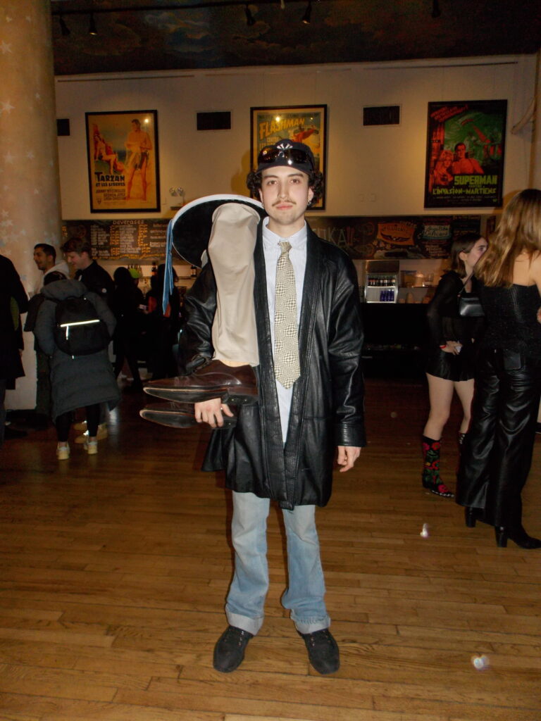 A man stands in the lobby with a side table over his shoulder, fashioned with dress pants and shoes on the table legs to imitate the lower-half of a body.