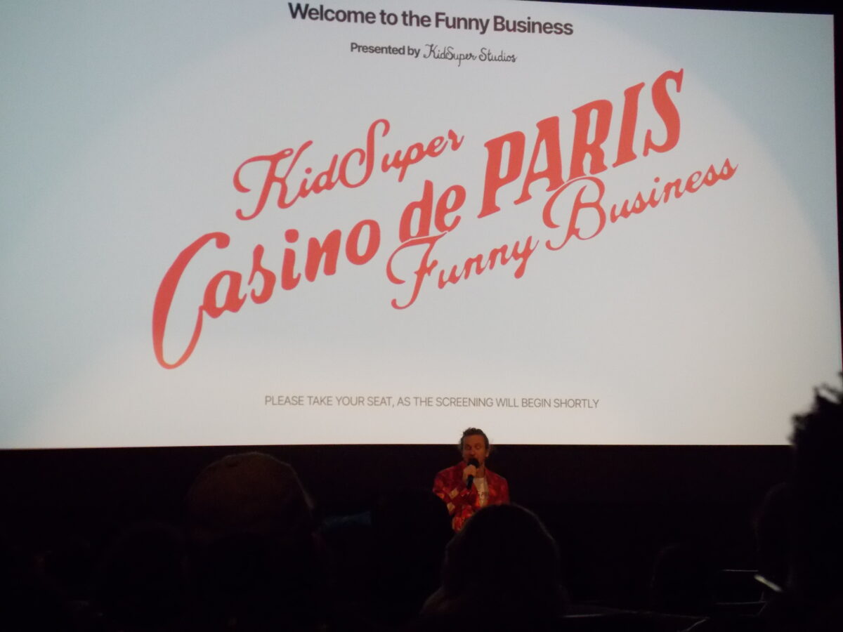 Colm Dillane, in a red suit and holding a microphone, stands in front of the theaters’ screen, which reads “KidSuper, Casino de Paris, Funny Business,” in red lettering against a white background.