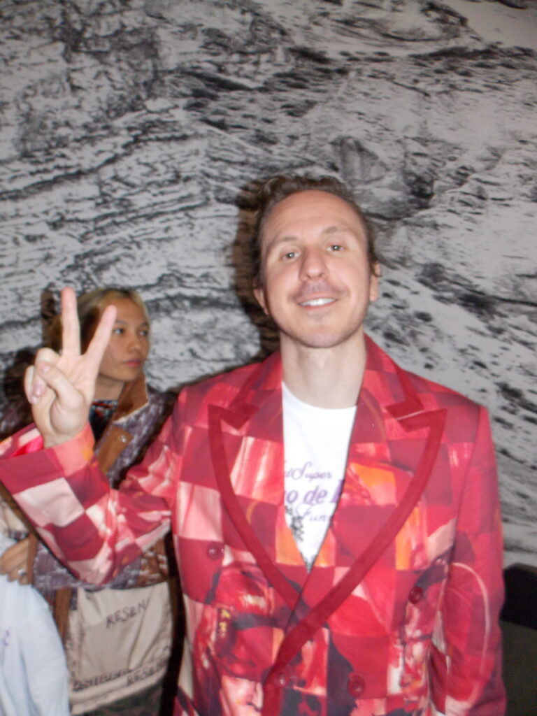 Colm Dillane poses for the camera, holding up a peace sign and wearing his own red patterned suit.