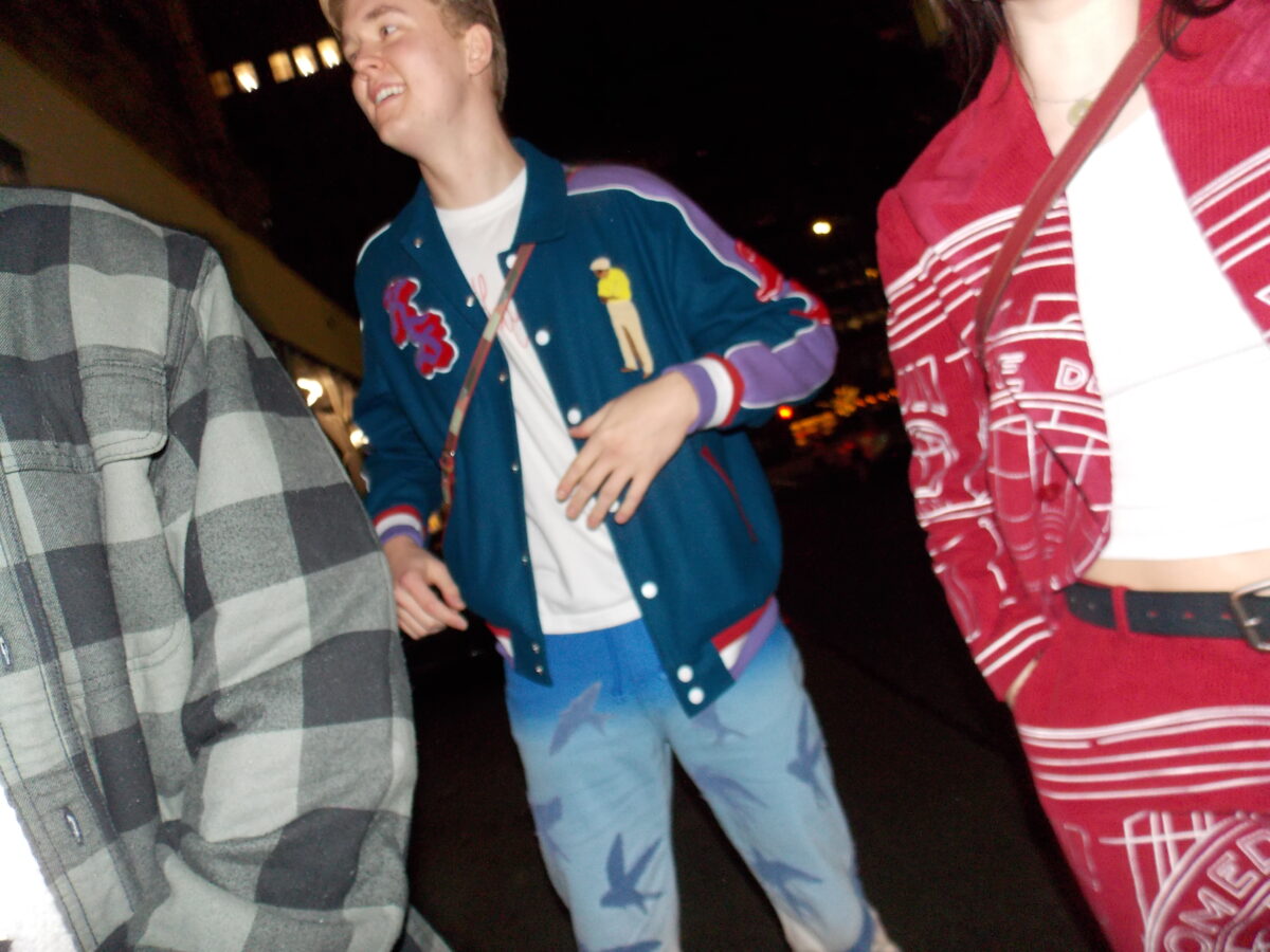 Photo of two people's torsos. One wearing a dark blue letterman jacket with fabric appliques and blue jeans with a large bird print, the other wearing a red suit with white stitching across the front.