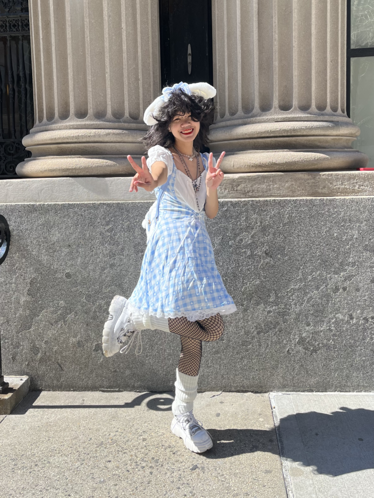 First-year illustration student Max Yuswardy wears a blue and white gingham dress with white platform sneakers and fuzzy white bunny ears while standing in front of two large stone columns.