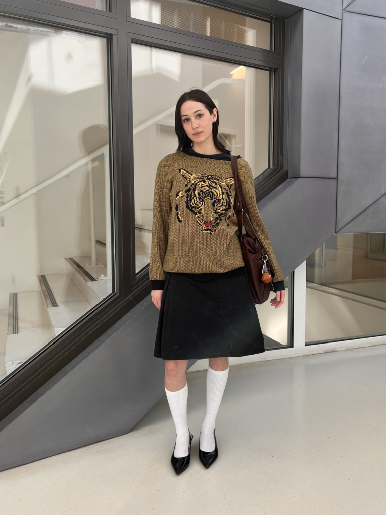 Second-year strategic design and management student Justice Trohan wears a tiger-printed sweater with a black midi skirt, tall white socks, and black kitten heels while standing in front of a school building.
