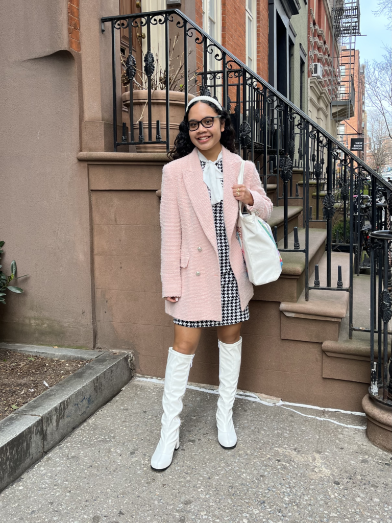 First-year journalism student Kea Humilde wears a black and white gingham dress, a pink tweed jacket, white knee-high boots, and a pearl headband while standing in front of a brownstone.