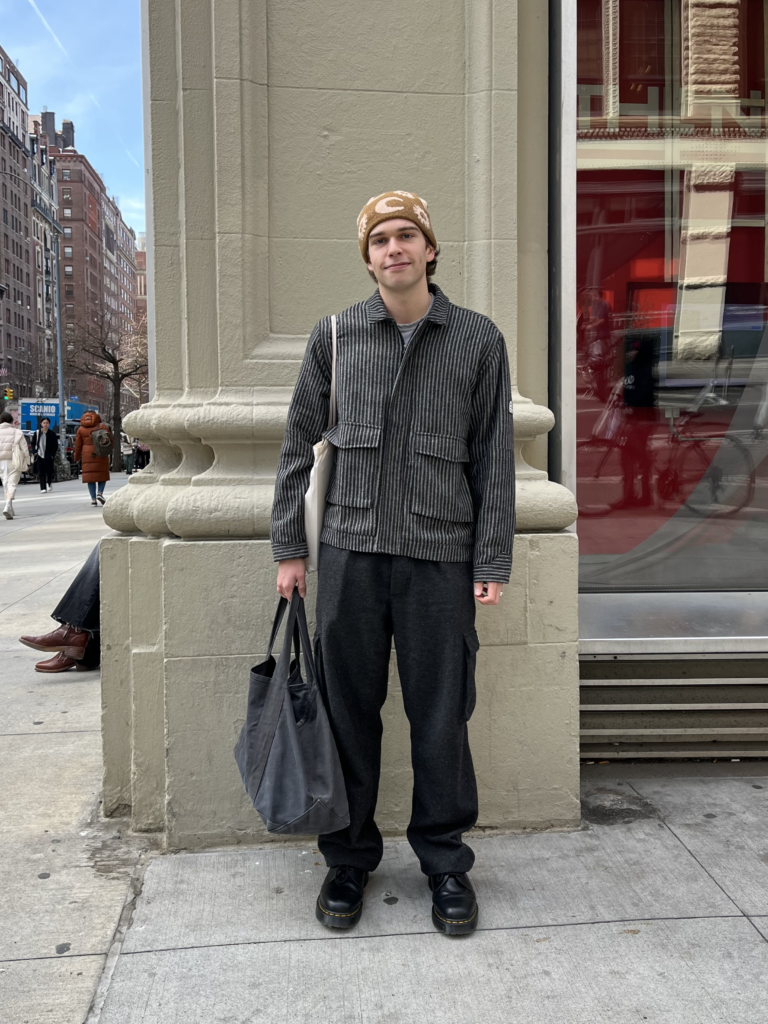 First-year philosophy student Stone Ferrand wears a great jacket, black pants, and a patterned beanie while standing in front of a school building.