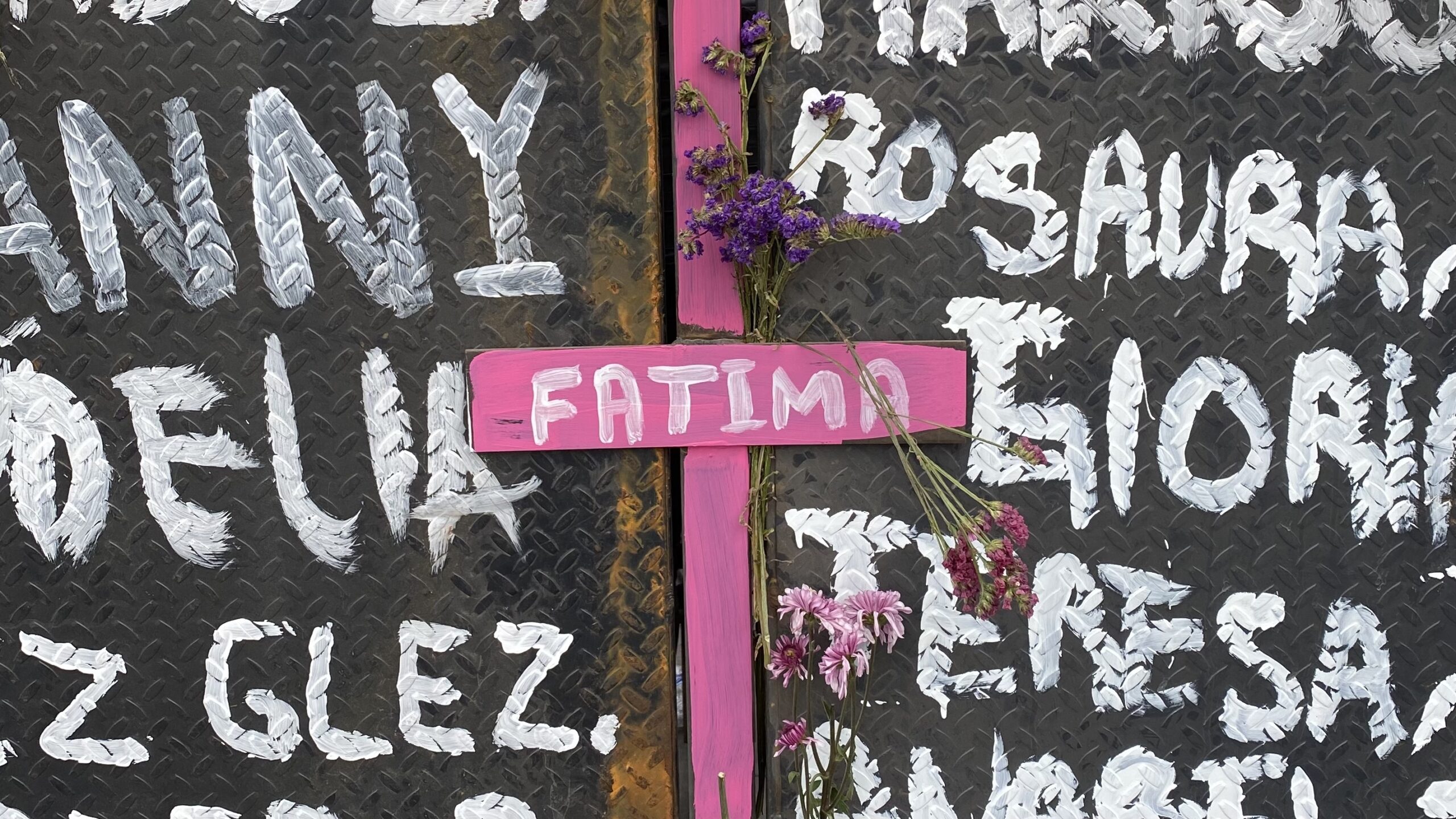 Pink cross with the name “Fatima” written with flowers hanging. The cross is hung on a black background and white names written around it.