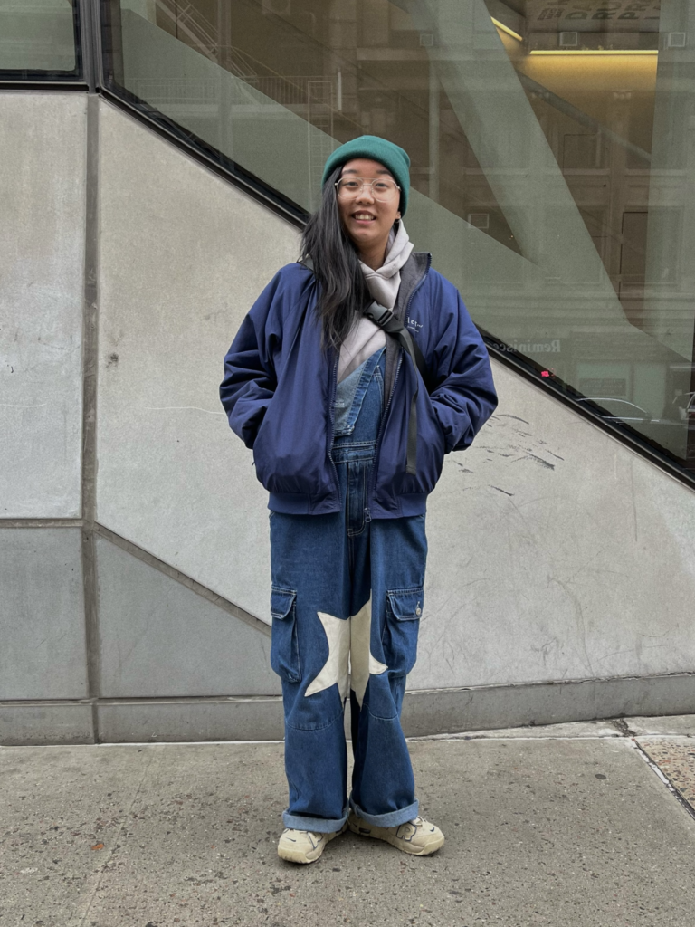 Art school student Emily Li stands on Fifth Avenue wearing denim overalls with star patches on the knees, a blue windbreaker jacket, and a green beanie.