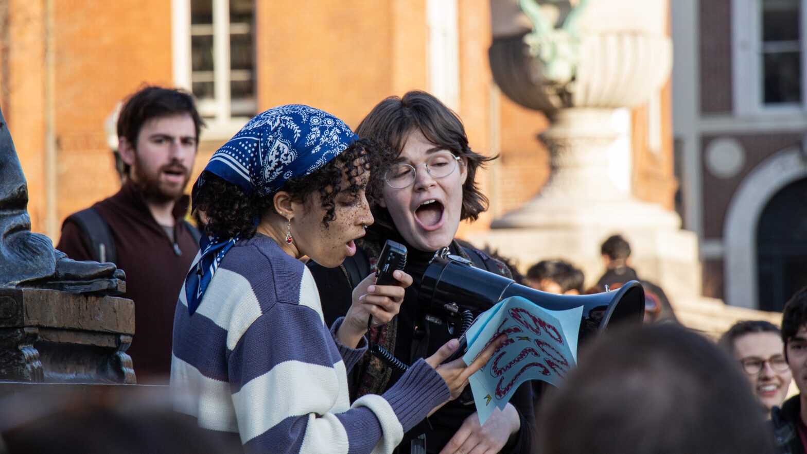 Two young women passionately speak into a bullhorn to a crowd. The one on the left has curly black hair tied back by a blue and white bandana, and is wearing a blue and white striped sweater. The woman on the right has short brown hair and a backpack on.