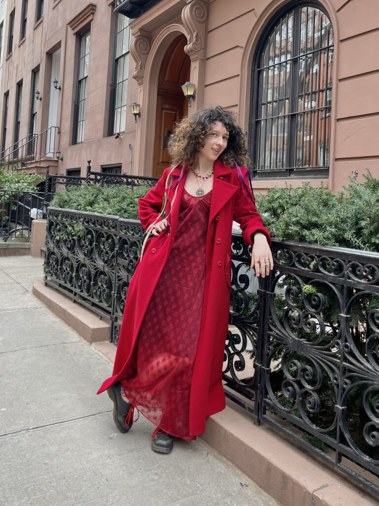 Art school student Innana Rose leans on a black railing in front of bushes and a brownstone while wearing a bright red coat with a matching red dress. 