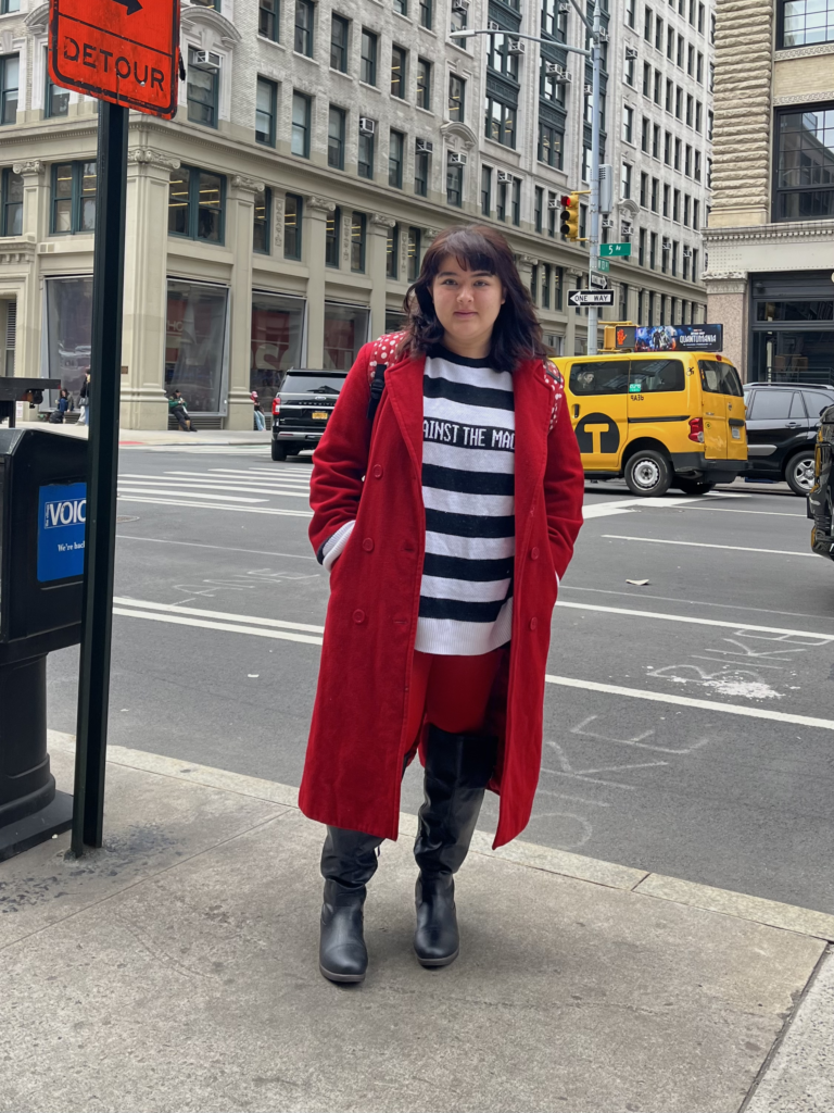 Art school student Kat Yoakum stands on Fifth Avenue wearing a red coat, black and white striped sweater, red tights, and black knee high boots.
